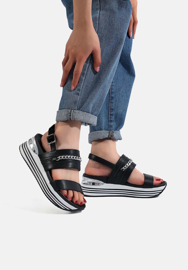 Sandali neri con cinturini e una larga piattaforma a righe bianche e nere. Presentano dettagli in catena metallica e una cinghia alla caviglia con fibbia.