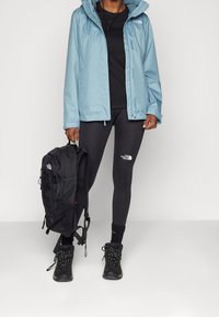 Vaaleansininen vedenpitävä takki, mustat leggingsit logolla, musta reppu ja mustat vaelluskengät nauhoilla, seisomassa valkoista taustaa vasten.