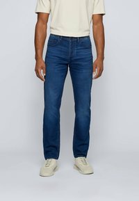 Man som bär mörkblå jeans, beige t-shirt och vita sneakers som står mot en enkel grå bakgrund.