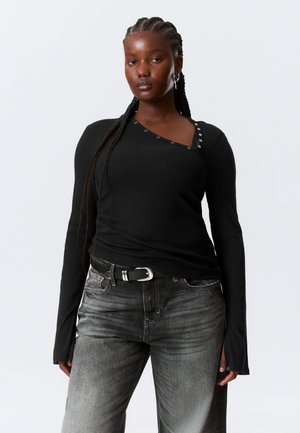 Femme aux cheveux tressés portant un haut noir asymétrique à manches longues avec boutons-pression et un jean noir délavé avec ceinture, debout face à l'avant.