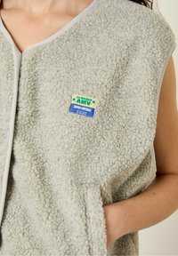 Svetr z fleece materiálu v světle šedé barvě s texturovaným povrchem. Obsahuje výstřih do V, otevřené strany a modrožlutou nášivku na přední straně.