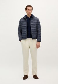 Grijze pufferjack met horizontale quiltstructuur, marineblauwe hoodie eronder, lichtbeige broek en bruine instappers.