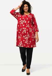 Rote Tunika mit Blumenmuster, drei Viertelarm, lockerer Schnitt, kombiniert mit schwarzen, engen Hosen und schwarzen Schuhen. Glatte Textur.