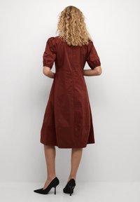 Ruda spalvos suknelė su pūkuotomis rankovėmis, iki kelių ilgio, su prigludusia liemenėle. Dėvima su juodais aukštakulniais, lygia audinio tekstūra.