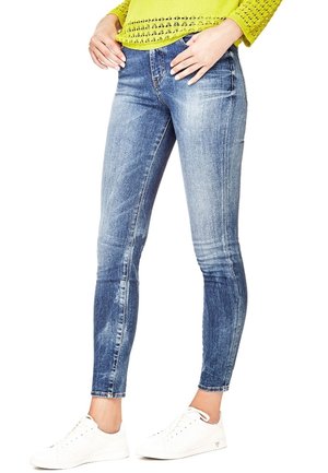 Frau trägt ausgewaschene blaue Skinny-Jeans mit weißen Sneakers und einen limettengrünen, gehäkelten Pullover, Hände ruhen in der Nähe der Taschen.