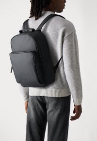 Schwarzer Leder-Rucksack mit rechteckiger Form, vorderer Reißverschlusstasche und gepolsterten Schulterträgern, getragen über einem hellgrauen Pullover.