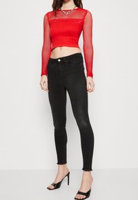 Mujer con un top corto de malla roja de manga larga, jeans ajustados de talle alto negros y zapatos de tacón negros abiertos, de pie contra un fondo blanco.