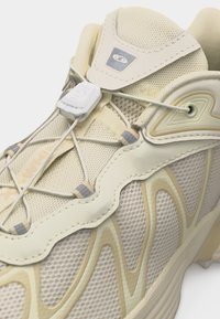 Κοντινό πλάνο μπεζ αθλητικού παπουτσιού με υφασμάτινο δίχτυ, επενδύσεις με κυματιστό σχέδιο, σύστημα γρήγορου δέσιμου και ταμπελάκι με το εμπορικό σήμα στη γλώσσα του παπουτσιού.