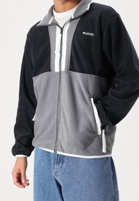 Fleecejacka med svarta och grå paneler, dragkedja fram och vita detaljer. Har två sidofickor och en ståkrage.