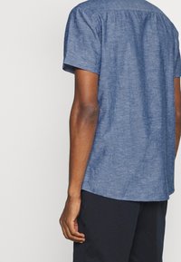 Camicia azzurra in lino a maniche corte con scollatura rotonda, vestibilità morbida e una leggera trama. La parte posteriore presenta una cucitura a yoke e un orlo curvo.