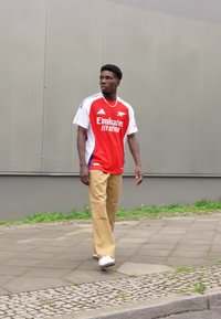 Maillot de sport rouge et blanc avec le logo "Emirates", associé à un pantalon ample beige et des baskets blanches. Cadre extérieur avec un fond gris.