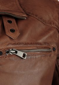 Gros plan sur une veste en cuir marron, avec une poche zippée en argent, des boutons-pression et des accents texturés. Surface en cuir lisse et perforée.