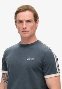 Camiseta de algodón azul marino de manga corta, con franjas de acento en crema y un logo bordado en el pecho. Textura suave y corte relajado.