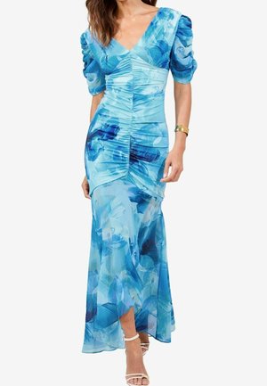 Kvinde iført en lang blå blomsterkjole med rynket front, puffede ærmer, V-hals og hvide sandaler med stropper, stående foran hvid baggrund.