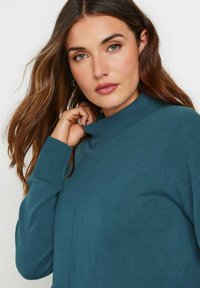 Maglione teal a collo alto con un colletto a costine e una texture liscia, con un design aderente e sottili dettagli delle cuciture sulla parte anteriore.