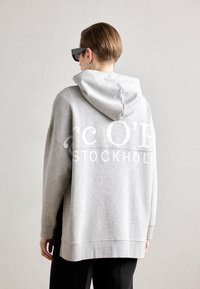 Hanorac oversized gri, prezentând o imprimare mare albă cu textul "rc O'H STOCKHOLM" pe spate, echipat cu glugă și manșete ribbate.