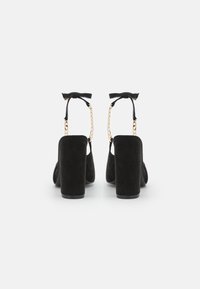 Chaussures à talons hauts en suede noir avec un talon épais, des brides de cheville et des accents en chaîne dorée. Elles présentent un détail de nœud à l'arrière.