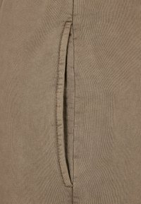 Beige stoff med en jevn tekstur som har en sidelomme. Sømdetaljer er synlige langs kanten av lommen.