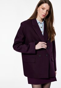 Blazer cruzado de tela púrpura oscuro, con botones negros, hombros estructurados y dos bolsillos delanteros. Se lleva sobre una camisa azul con cuello.