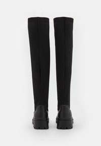 Polvisteiset mustat saappaat, jotka on valmistettu joustavasta kankaasta tekstuuripinnalla; ominaisuuksiin kuuluvat tukevat kumipohjat ja ribbikudotut yläosat.