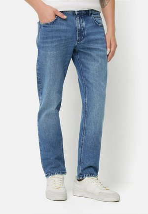 Mand iført blå jeans med lige ben og hvide sneakers, stående med den ene hånd i lommen på en ensfarvet baggrund.
