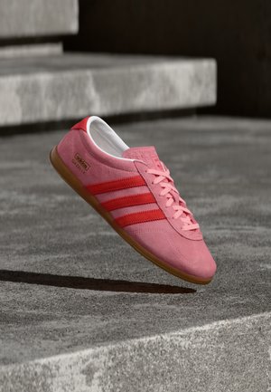 Zapatilla de ante rosa con rayas rojas, suela de goma y cordones rosas. Presenta el logo "Adidas Gazelle" en el lateral.