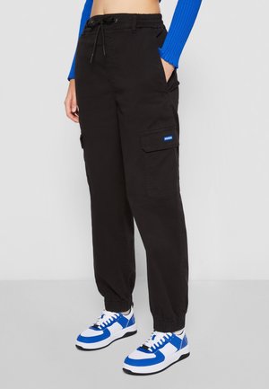 Black cargo pants with elastic waistband, drawstring, side pockets, and blue logo patch. Worn with blue and white sneakers.