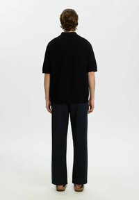 Personne debout, vue de dos, portant une chemise noire à manches courtes, un pantalon noir et des chaussures sans lacets, sur un fond blanc uni.