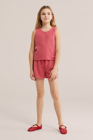 Young girl standing wearing sleeveless pink knit top, matching shorts, and red woven Mary Jane shoes against neutral background.