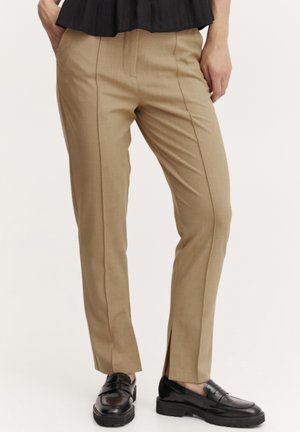 Slim-fit béžové kalhoty s hladkým povrchem, vertikálními švy a bočními rozparky na lemu, v kombinaci s černými mokasíny.