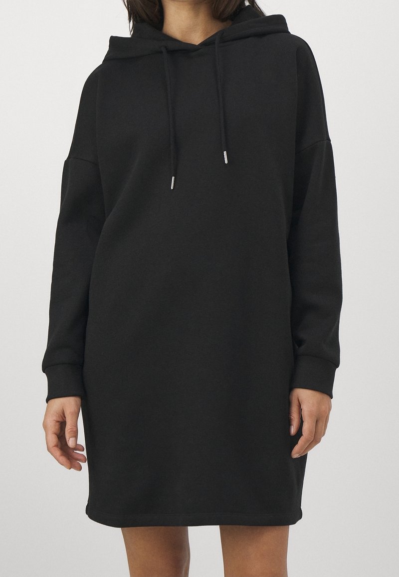 Femme portant une robe sweat à capuche noire ample avec manches longues et cordons, debout devant un fond clair uni.