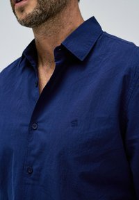 Camisa de botones azul marino en tejido ligero, que cuenta con un cuello clásico y un pequeño logotipo bordado en el lado izquierdo.