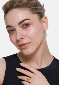 Goldring mit Sonnenstrahlen-Design und zentralem Stein. Das Model hat helle Haut, blaue Augen und trägt ein ärmelloses dunkles Oberteil. Glatte Hautstruktur.