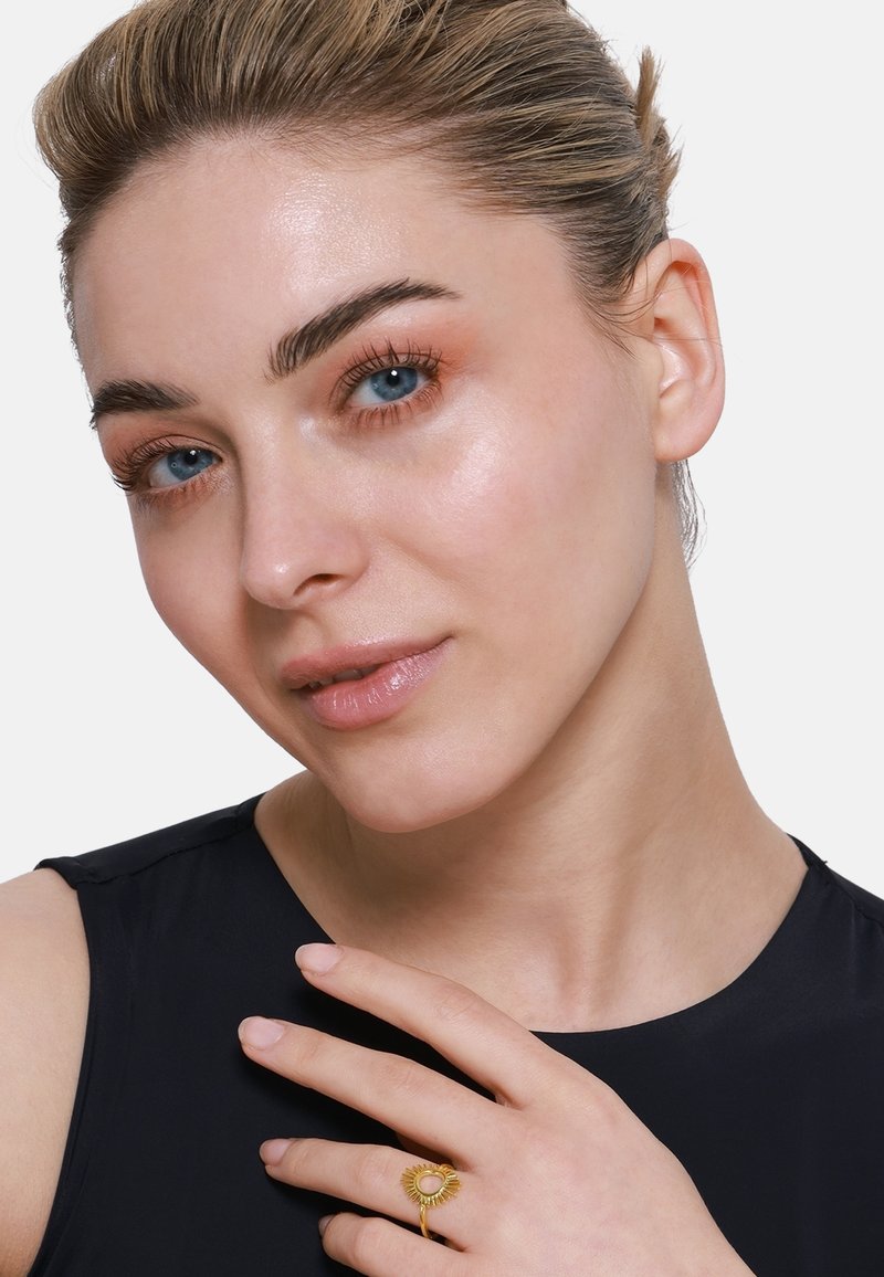 Goldring mit Sonnenstrahlen-Design und zentralem Stein. Das Model hat helle Haut, blaue Augen und trägt ein ärmelloses dunkles Oberteil. Glatte Hautstruktur.