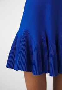 Ourlet de jupe plissée bleue avec détail motif zigzag porté au-dessus du genou sur une personne devant un fond uni.
