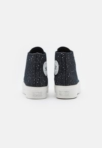 Svarta högsnörade sneakers i canvas med en fläckig textur, vit gummisula och cirkulär logopatch på hälen. Ren design, minimala accenter.