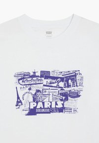 Valkoinen puuvillapaita, jossa on sininen graafinen kollaasikuva pariisilaisista kyltistä, maamerkkejä ja sana "BOULANGERIE" korostettuna.