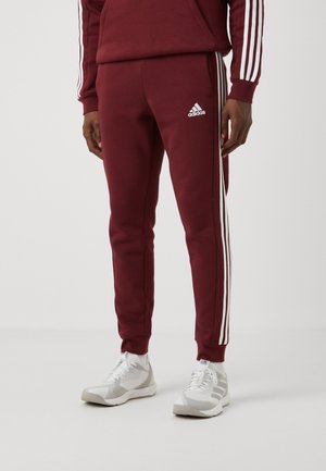 Mann trägt bordeauxrote Adidas-Trainingshose mit weißen Streifen und weiße Turnschuhe, steht vor einem einfarbigen hellen Hintergrund.