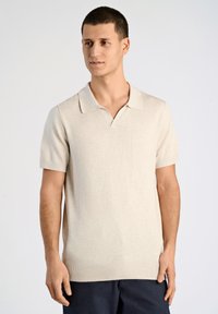 Ljus beige kortärmad polo med V-ringning, stickad textur och ribbat avslut. Har en klassisk kragdesign.