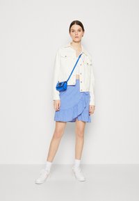 Veste en denim blanc sur un top court, associée à une jupe froncée à pois bleus. Baskets blanches avec des accents roses et un sac bandoulière bleu.