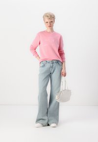 Pink sweatshirt with white logo, light blue flared jeans, and white sneakers. Holding a white handbag with a curved design.
