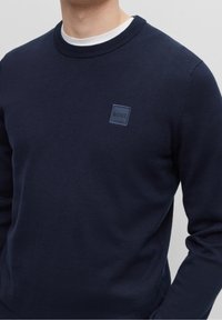 Suéter de punto azul marino con cuello redondo acanalado, que presenta un pequeño parche con logo cuadrado en la esquina superior izquierda. Textura suave.
