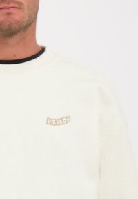 Sweatshirt en polaire crème avec un col rond, arborant une étiquette textuelle écrite « VOLCOM » en off-white cousue sur la poitrine gauche. Texture douce, coupe décontractée.