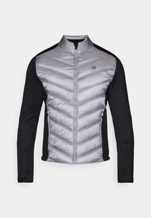 Padded jacket with a silver, quilted front and black knit sleeves. Features a high collar, zip front, and logo on the chest.