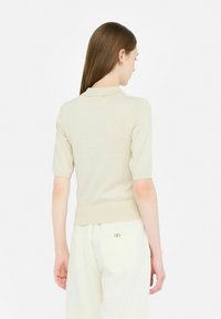 Maglione polo beige a maniche corte con texture a costine e design aderente. Indossato con pantaloni bianchi a vita alta con un accento dorato sulla tasca.