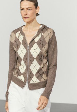 Femme portant un sweat à capuche marron à fermeture éclair avec motif argyle et un pantalon blanc, debout devant un fond clair uni.