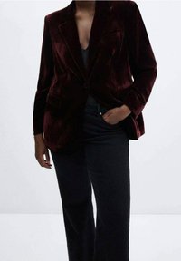 Personne portant un blazer en velours bordeaux foncé avec un bouton, un haut noir et un pantalon noir ample avec une main dans la poche.