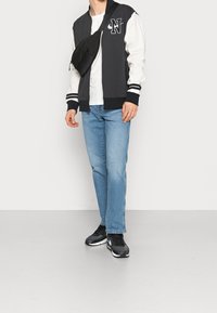 Svart och vit bomberjacka med randiga manschetter, vit t-shirt, blå jeans och svarta sneakers med vita accenter. Svart axelväska.