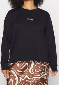 Sudadera negra de manga larga y cuello redondo, con un logo blanco de "Calvin Klein" en el centro. Tejido suave, corte relajado.