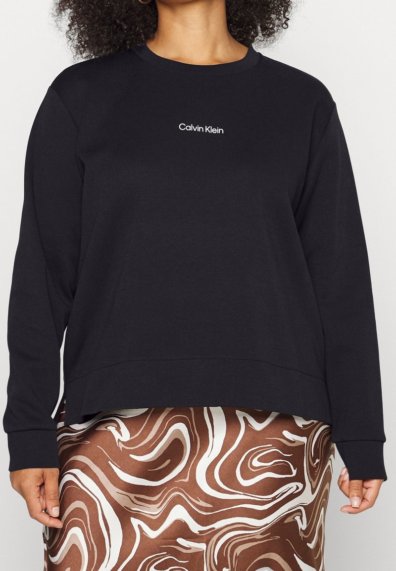 Sudadera negra de manga larga y cuello redondo, con un logo blanco de "Calvin Klein" en el centro. Tejido suave, corte relajado.